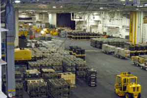 Ein großes Lagerhaus voller Numerous Trucks, Container, Menschen, Lichter, Stangen und andere Gegenstände mit einem prominenten Rad.