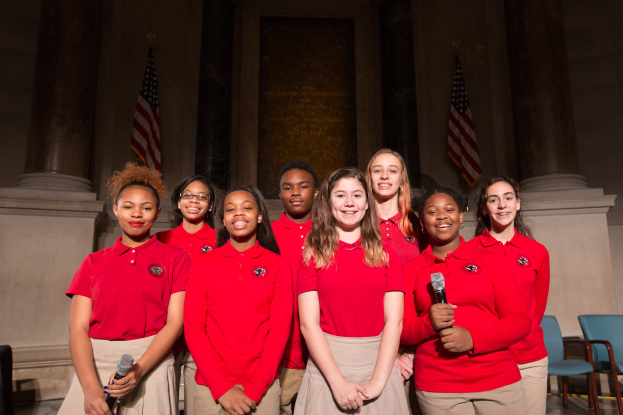 Eine Gruppe junger Frauen in roten Hemden, die zusammenstehen, lächeln und Mikrofone halten, mit Stühlen, Säulen, Fahnen und einer Wand im Hintergrund.