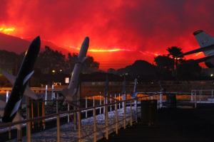 Ein Flugzeug fliegt über eine Start- und Landebahn mit Raketen, umgeben von Zäunen, Mülltonnen, Bäumen, Laternenmasten und Gebäuden, mit von den Kalifornien-Wildfires verqualmten Himmel im Hintergrund.