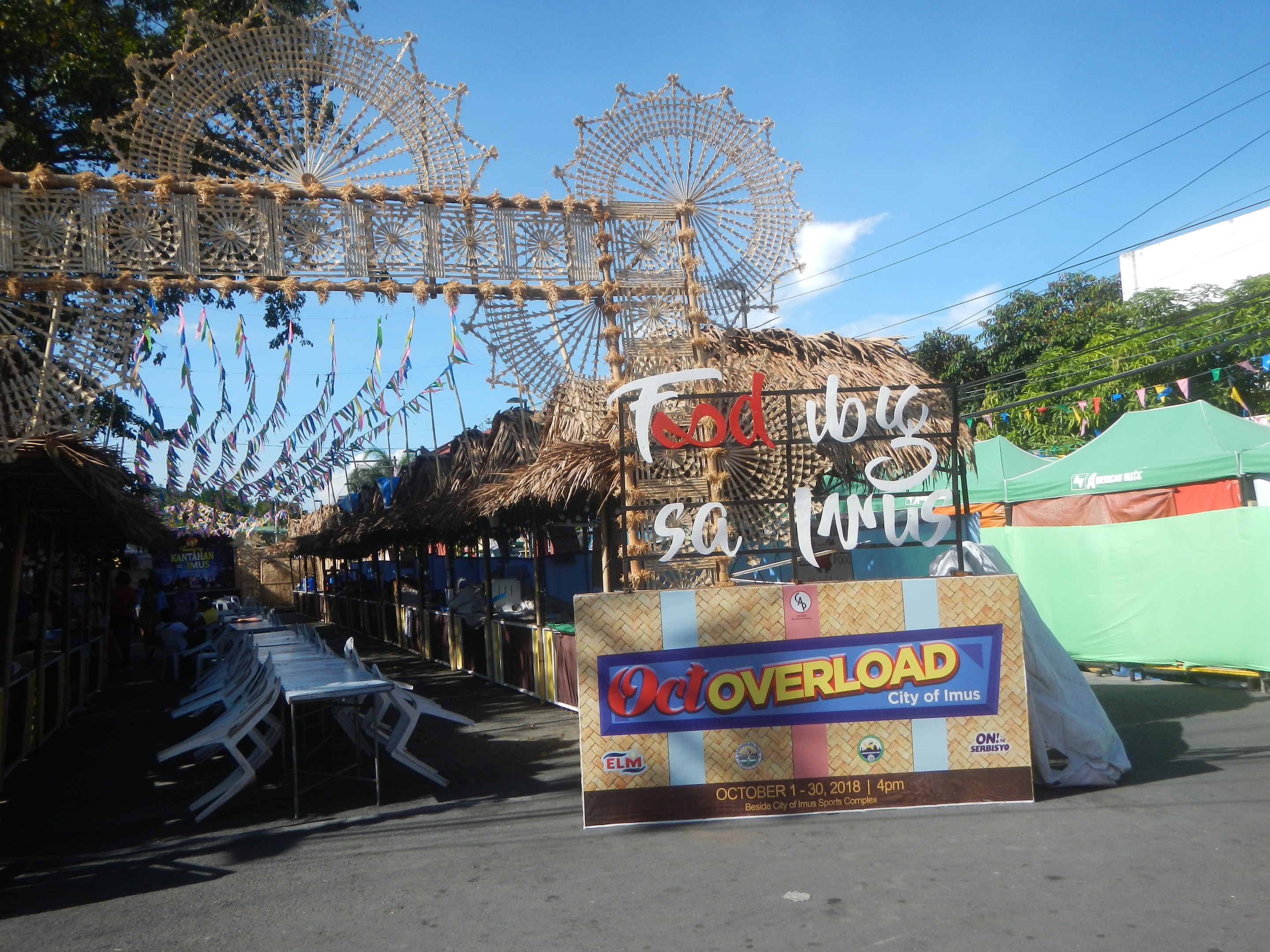 Eine Karnevalsszene mit einem großen Riesenrad in der Mitte, umgeben von Hütten, Sitzbereichen, einem Schild, Dekorationen, Bäumen, Überlandleitungen und einem bewölkten Himmel.