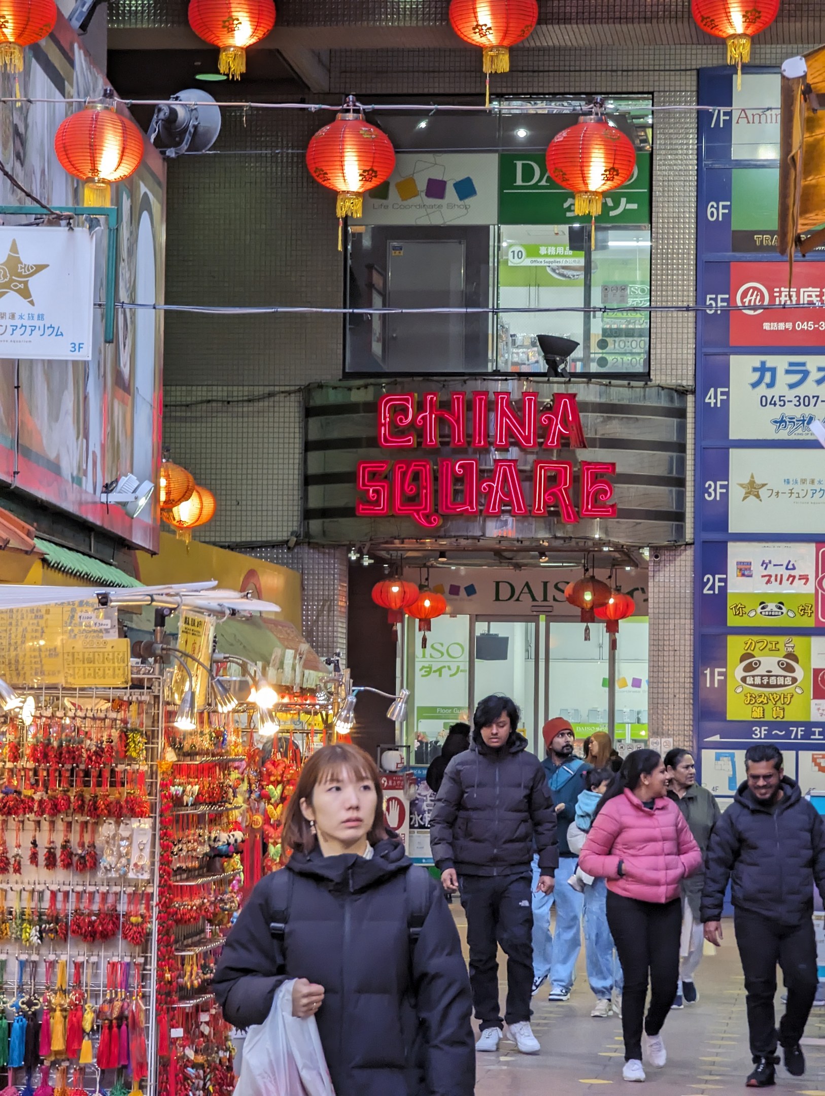 Eine Gruppe von Menschen geht eine Straße mit Geschäften auf beiden Seiten entlang, wobei eine Frau mit einer Tasche im Vordergrund zu sehen ist. Auf der linken Seite des Bildes befinden sich verschiedene Verkaufsartikel, und im Hintergrund ist ein Gebäude mit einem Namensschild und roten Laternen zu sehen.