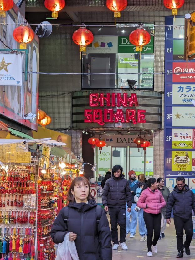 Eine Gruppe von Menschen geht eine Straße mit Geschäften auf beiden Seiten entlang, wobei eine Frau mit einer Tasche im Vordergrund zu sehen ist. Auf der linken Seite des Bildes befinden sich verschiedene Verkaufsartikel, und im Hintergrund ist ein Gebäude mit einem Namensschild und roten Laternen zu sehen.