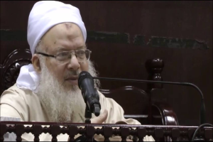 Asaduddin Owaisi, ein Mann mit einem weißen Bart und Turban, spricht in ein Mikrofon vor einer hölzernen Balustrade, bekleidet mit einem cremefarbenen Anzug und Brille, vor einem dunklen Hintergrund.