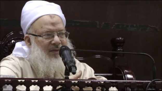 Asaduddin Owaisi, ein Mann mit einem weißen Bart und Turban, spricht in ein Mikrofon vor einer hölzernen Balustrade, bekleidet mit einem cremefarbenen Anzug und Brille, vor einem dunklen Hintergrund.