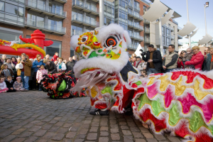 Ein lebhaftes chinesisches Neujahrsfest in Amsterdam mit einer Löwen-Tanzvorstellung vor einer Zuschauermenge, einige halten Kameras, vor einem Hintergrund aus Gebäuden, Laternenmasten und einem klaren blauen Himmel.