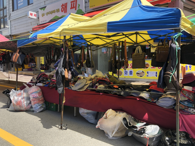 Ein Straßenmarkt mit Zelten, die Taschen, Hüte und andere Gegenstände präsentieren, vor Gebäuden mit Fenstern und Texttafeln.
