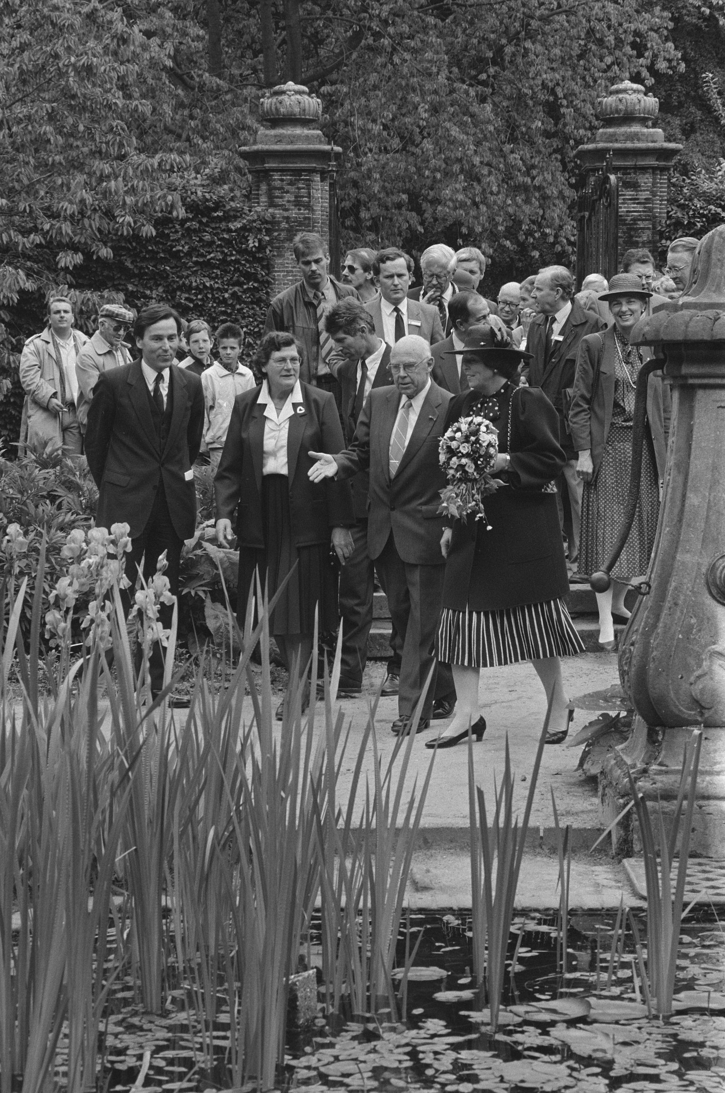Schwarzes und weißes Bild einer Gruppe von Menschen, die sich um einen Teich in einem Garten versammelt haben, mit einer Frau in der Mitte, die einen Blumenstrauß hält, und einem Mann in einem Anzug rechts von ihr, umgeben von üppigen Pflanzen, Bäumen und Säulen.