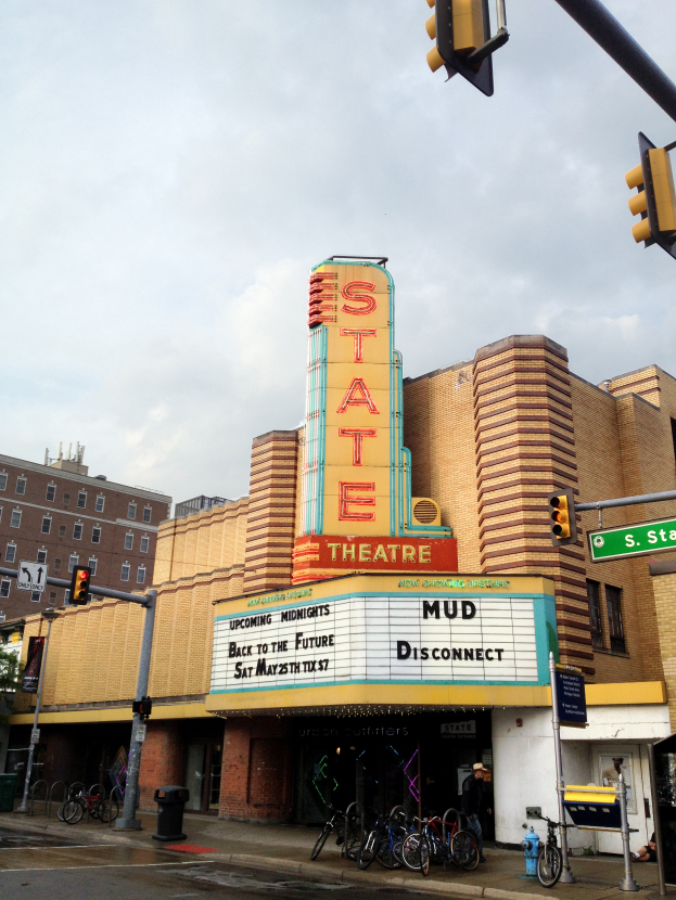 Das Staatstheater in Detroit, Michigan, mit einer Schautafel, Mülltonne, parkenden Fahrrädern, einer Person, Pfosten, Ampeln, Schautafeln, einem Baum und einem bewölkten Himmel.