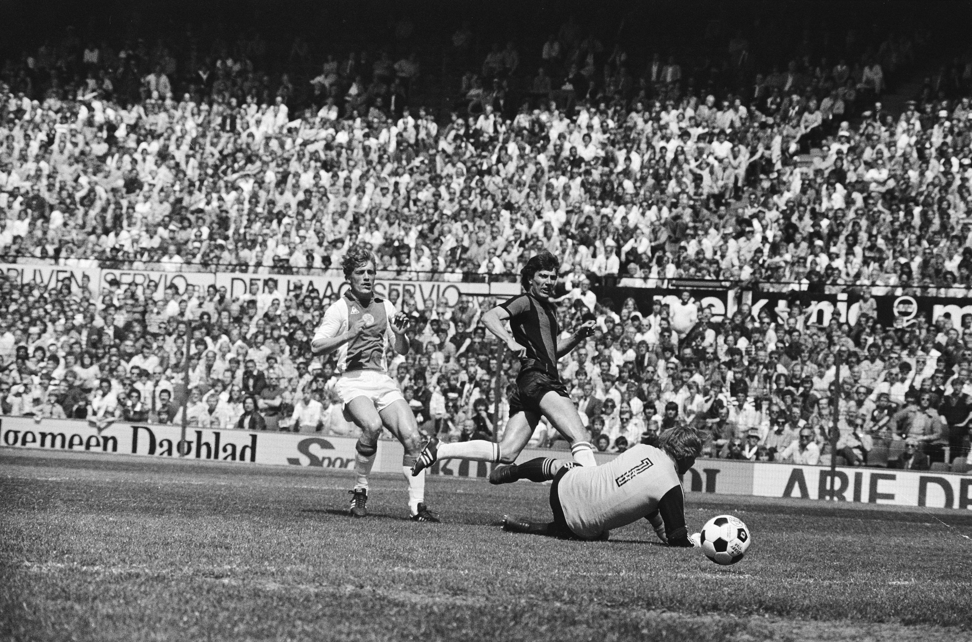 Schwarz-weißes Foto von Männern, die auf einem Feld Fußball spielen, mit Zuschauern im Hintergrund und einer Bundesliga-Banner von 1978-79 Ajax 1-1 Hannover 96.