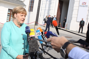 Bundeskanzlerin Angela Merkel spricht vor einem Gebäude mit Fenstern und einer Tür zu den Medien, umgeben von einer Gruppe von Menschen, wobei eine Person ein Mobiltelefon und eine andere eine Uhr trägt.