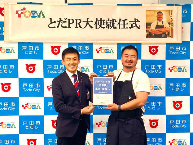 Zwei lächelnde Männer mit einem Zertifikat, mit einem Banner im Hintergrund, das "Toda City Wins PR Award" lautet und ein Foto einer Person zeigt.