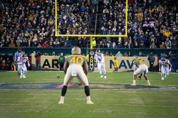 Fußballspieler mit Helmen und Sportbekleidung konkurrieren auf einem Feld mit Zuschauern im Hintergrund, Stadionstrukturen und sichtbare Beschilderung.