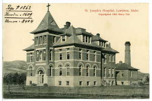 Ein Schwarzweiß-Foto des St. Joseph-Krankenhauses in Lewiston, Idaho, das ein großes Gebäude mit Fenstern, eine Treppe mit Geländern, einen Schornstein auf der rechten Seite und Text oben zeigt, mit Bäumen, Hügeln und Himmel im Hintergrund.