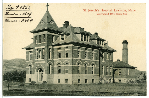 Ein Schwarzweiß-Foto des St. Joseph-Krankenhauses in Lewiston, Idaho, das ein großes Gebäude mit Fenstern, eine Treppe mit Geländern, einen Schornstein auf der rechten Seite und Text oben zeigt, mit Bäumen, Hügeln und Himmel im Hintergrund.