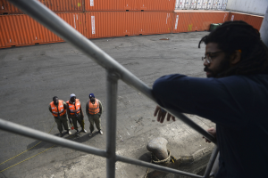Ein Mann in einer dunklen Jacke steht auf einem Metallgelönder in der Nähe einer Gruppe von Menschen in orangefarbenen Jacken und Kappen, mit Containern und einer Wand im Hintergrund am Hafen von Los Angeles.