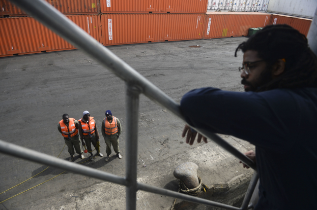Ein Mann in einer dunklen Jacke steht auf einem Metallgelönder in der Nähe einer Gruppe von Menschen in orangefarbenen Jacken und Kappen, mit Containern und einer Wand im Hintergrund am Hafen von Los Angeles.