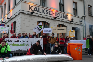 Eine Gruppe von Menschen mit Schildern und Plakaten steht vor einem Gebäude, mit zwei Personen im Vordergrund und einem Müllcontainer auf der rechten Seite, während einer Demonstration in Deutschland.