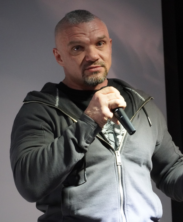 Ein Mann in einer schwarzen Kapuzenjacke und einem schwarzen Anzug mit einem weißen Hemd, glattem Haar und einem selbstbewussten Gesichtsausdruck h├Ąlt ein Mikrofon, waehrend er vor einer Wand steht.