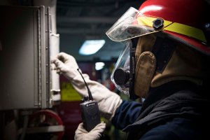 Ein Feuerwehrmann in voller Montur hält ein Funkgerät und ein metallisches Objekt, mit einem unscharfen Hintergrund.