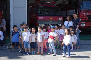 Eine Gruppe von Kindern steht vor einem Feuerwehrauto, einige tragen Mützen, mit mehreren anderen Feuerwehrautos im Hintergrund auf einem Feuerwehrausstellungsgelände.