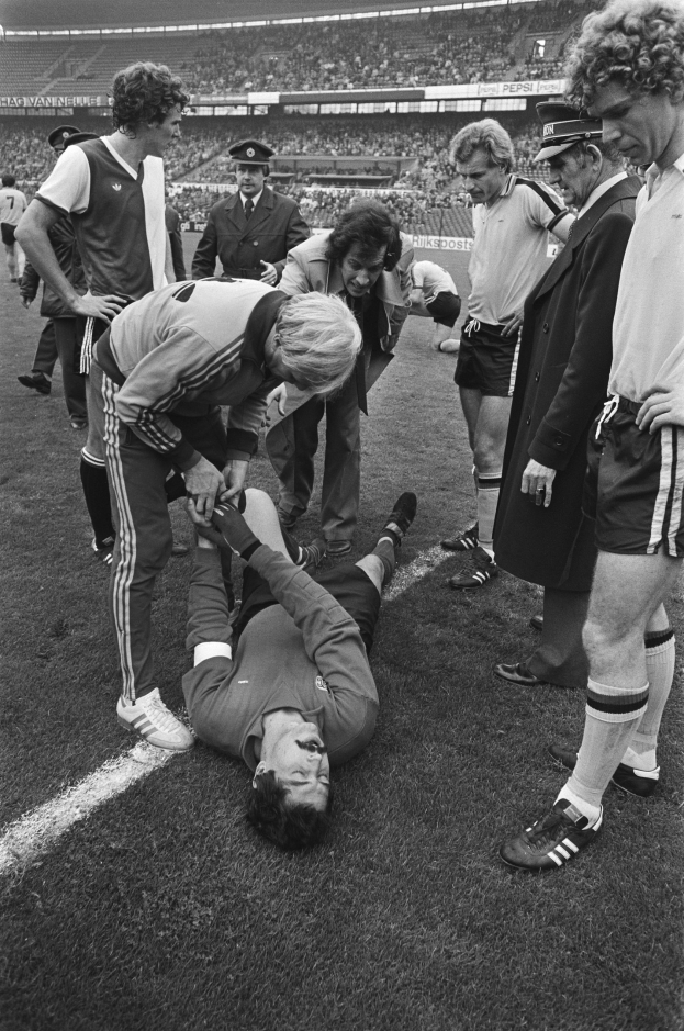 Schwarzes und weißes Bild von Fußballspielern und medizinischem Personal, das einen verletzten Spieler auf dem Feld betreut, während Zuschauer aus den Tribünen zuschauen.