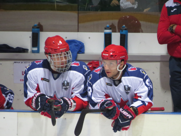 Gruppe junger Männer beim Eis-hockeyspielen in Sportkleidung, mit Eishockeyschlägern, Flaschen und Kleidung an einer Wand und einer Glasbarriere im Hintergrund.